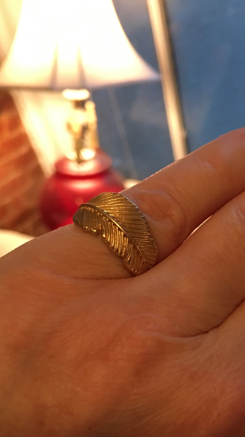 May include: A gold ring with a feather design. The ring is on a person's finger.