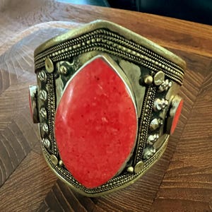May include: A wide, ornate cuff bracelet with a large, teardrop-shaped red stone at the center. The bracelet has a gold-colored metal base with intricate detailing, including braided and studded accents. Two smaller red stones are set on the sides.