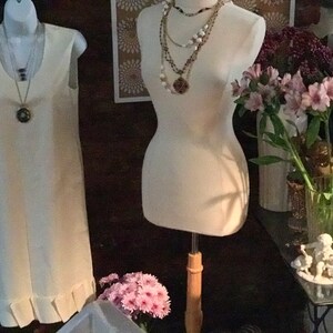 May include: Two white dress forms, one with a white sleeveless dress and the other with a layered necklace. The dress form on the left has a ruffled hem. The dress form on the right is wearing a layered necklace with a large pendant. The dress forms are in front of a dark brown wall with a floral patterned wallpaper behind the dress form on the right. There are pink flowers in a vase on the right side of the image.