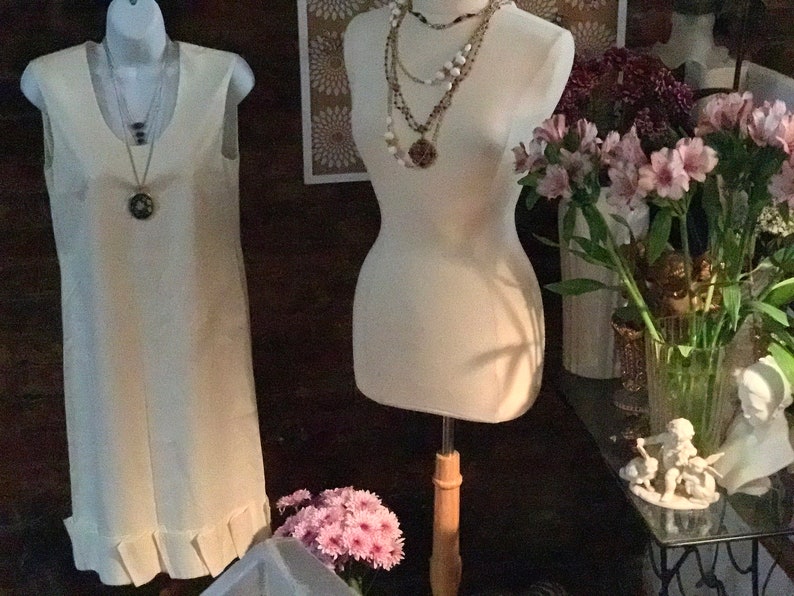 May include: Two white dress forms, one with a white sleeveless dress and the other with a layered necklace. The dress form on the left has a ruffled hem. The dress form on the right is wearing a layered necklace with a large pendant. The dress forms are in front of a dark brown wall with a floral patterned wallpaper behind the dress form on the right. There are pink flowers in a vase on the right side of the image.