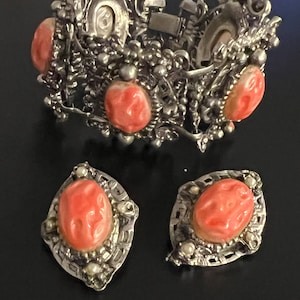 Op de afbeelding: Een vintage sieradenset met een armband en bijpassende oorbellen. De armband en oorbellen hebben een zilverkleurig metalen filigraanontwerp. Elk stuk heeft ovale, koraalkleurige stenen. De oorbellen zijn clip-on stijl.