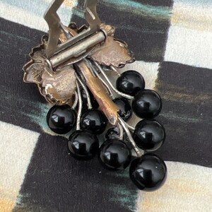 May include: A silver-toned brooch in the shape of a grapevine with black glass grapes. The brooch has a clasp on the back.