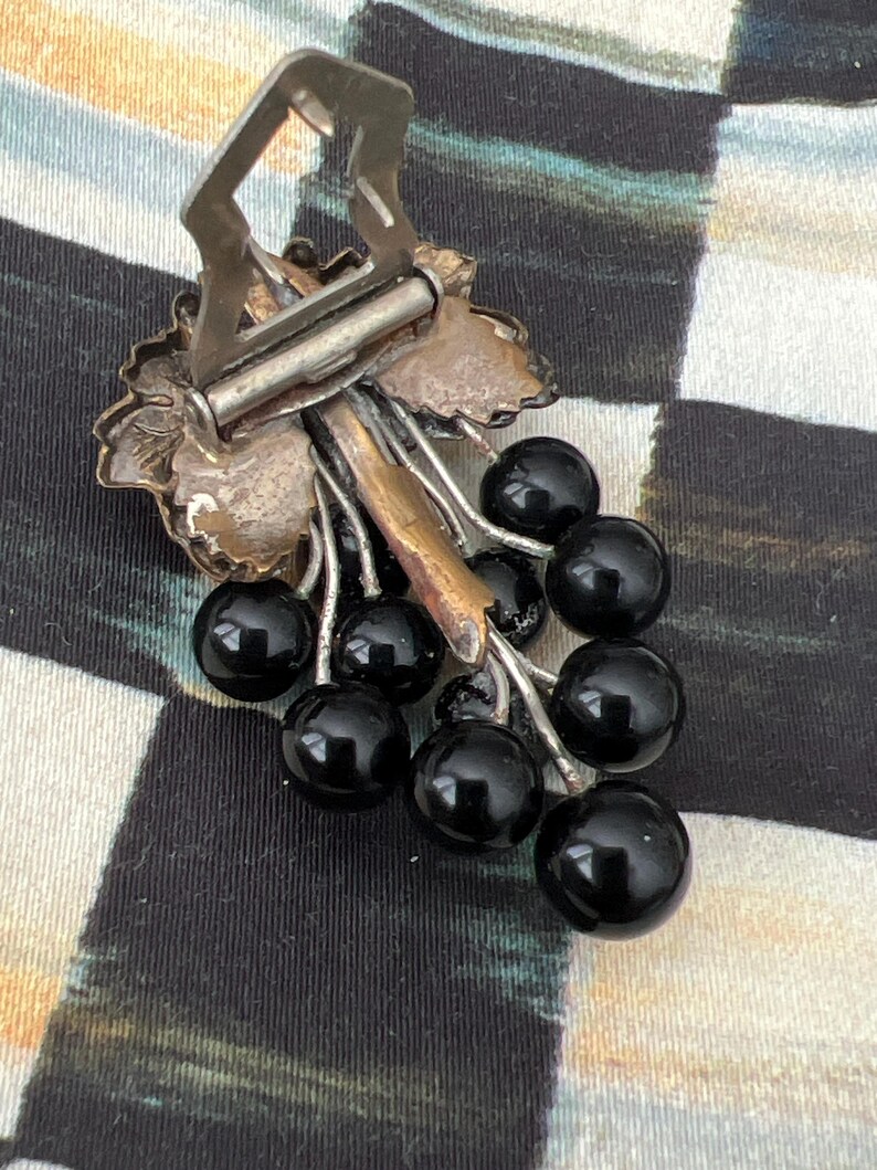 May include: A silver-toned brooch in the shape of a grapevine with black glass grapes. The brooch has a clasp on the back.