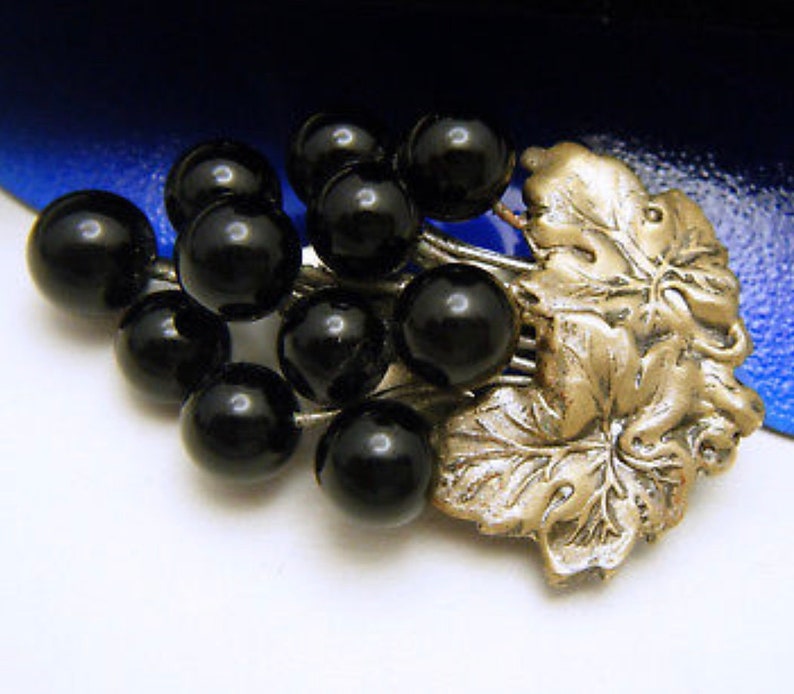 May include: A silver grapevine brooch with black glass grapes. The brooch is detailed with leaves and vines.