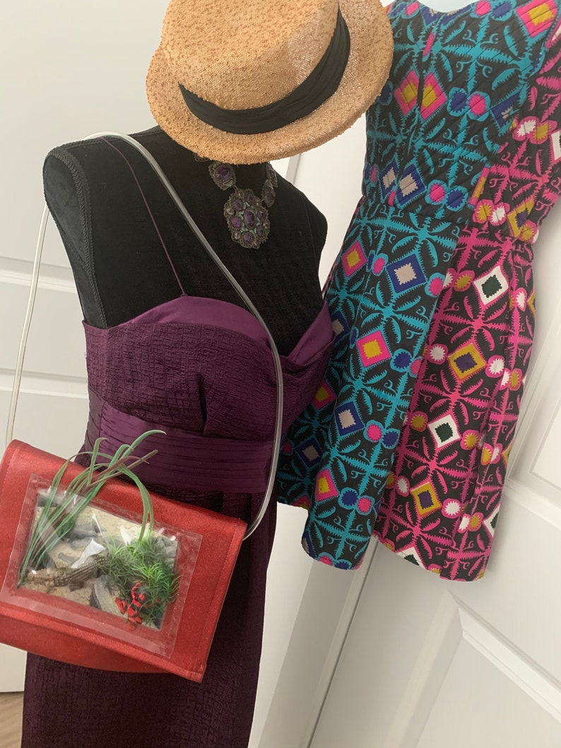 May include: A red purse with a clear plastic window showing a small potted plant inside. The purse is worn on a mannequin with a purple dress, a straw hat, and a large purple gemstone necklace.