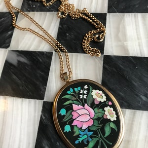 May include: A gold chain necklace with a black oval pendant featuring a colorful floral design. The pendant has a pink flower, blue and green leaves, and white flowers.