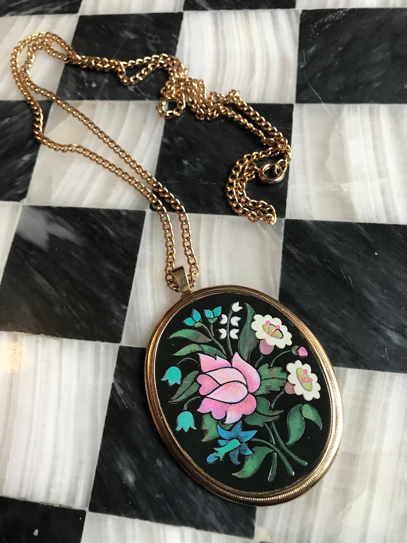 May include: A gold chain necklace with a black oval pendant featuring a colorful floral design. The pendant has a pink flower, blue and green leaves, and white flowers.