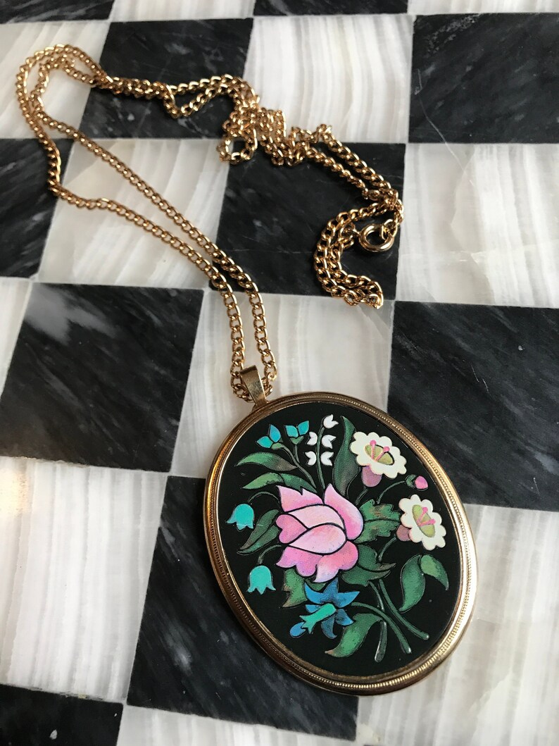 May include: A gold chain necklace with an oval pendant featuring a black background and a colorful floral design. The pendant has a gold rim and the flowers are pink, white, blue, and green.