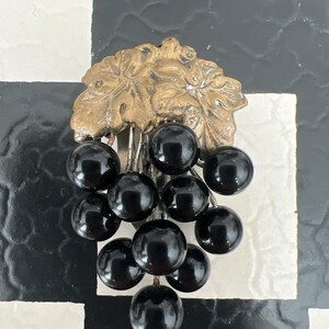 May include: A vintage brooch featuring a cluster of black glass grapes hanging from a gold-toned metal vine.