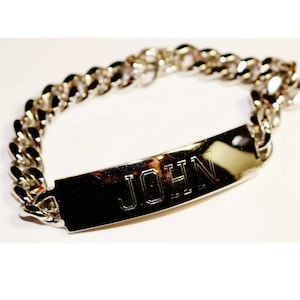 May include: A silver-colored curb chain bracelet with a rectangular nameplate. The nameplate is engraved with the name "JOHN" in capital letters. The bracelet is made of a shiny, metallic material.