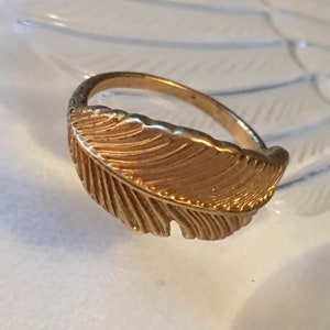 May include: A gold-toned ring with a feather design. The feather is detailed with lines and has a slightly curved shape.