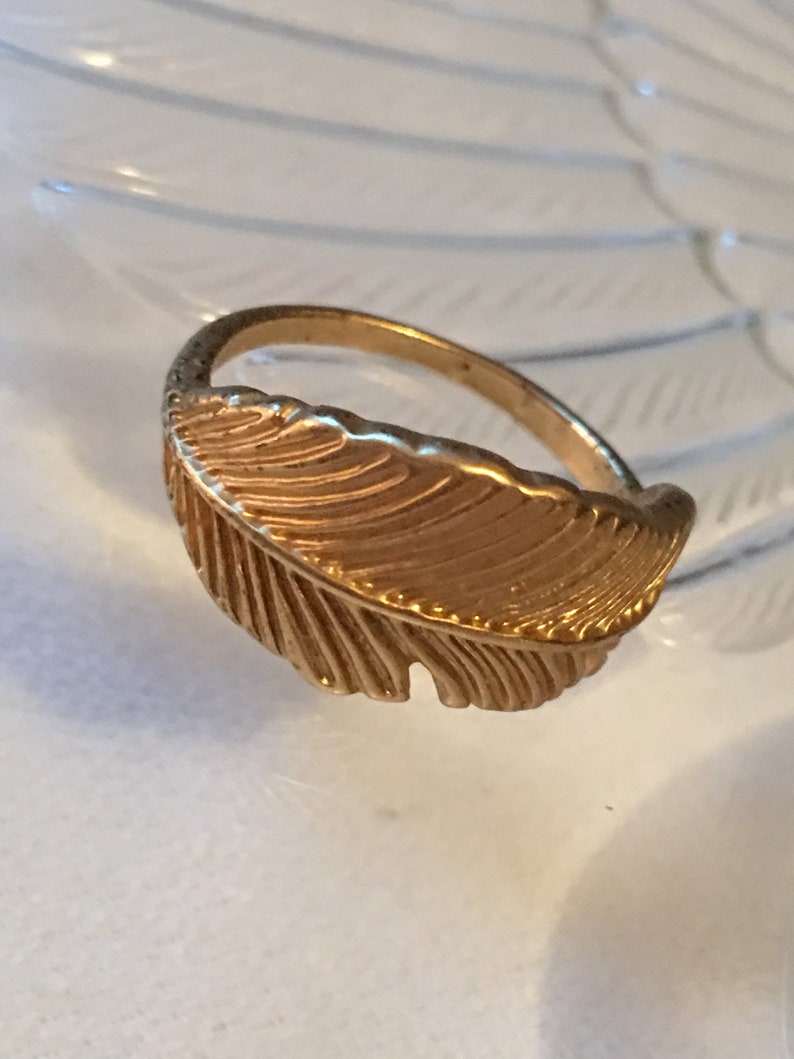 May include: A gold-toned ring with a feather design. The feather is detailed with lines and has a slightly curved shape.