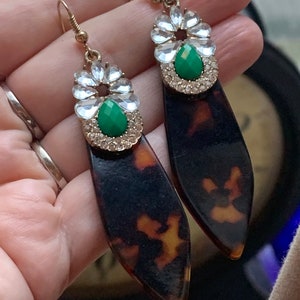 Puede incluir: Un par de pendientes colgantes con un diseño de caparazón de tortuga. Los pendientes presentan una piedra preciosa verde rodeada de diamantes de imitación transparentes.