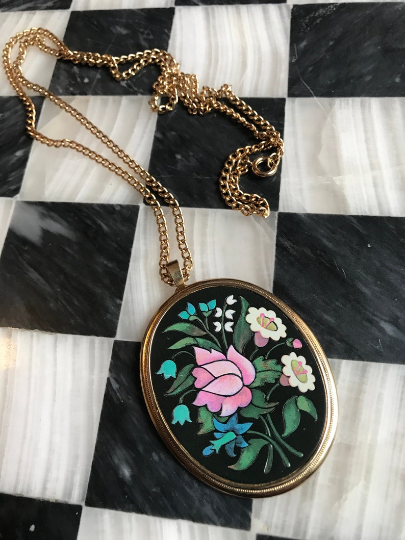 May include: A gold chain necklace with a black oval pendant featuring a colorful floral design. The pendant has a pink rose, white flowers, and blue and green leaves.