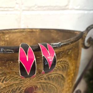 May include: A pair of hoop earrings with a black and pink geometric design. The earrings have a gold-colored metal frame and a glossy finish. The earrings are displayed on a gold-colored decorative bowl.