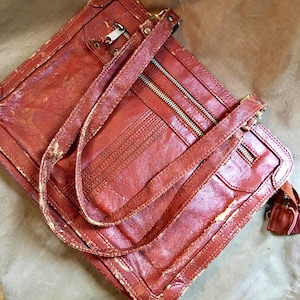Puede incluir: Un bolso vintage de cuero marrón con varios compartimentos con cremallera y una correa larga. El bolso tiene un aspecto desgastado y envejecido.