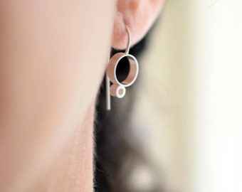May include: A close-up of a silver earring with a geometric design. The earring features a circular shape with a smaller circle and a straight bar. The earring is worn on an ear.