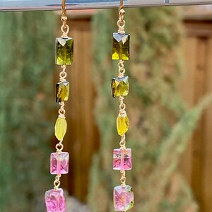 May include: A pair of gold earrings with a long, dangling design. The earrings feature multiple green and pink rectangular gemstones.