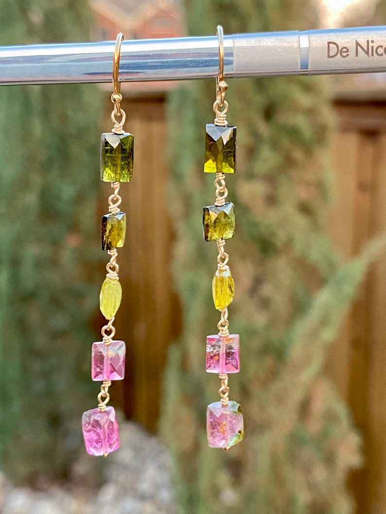 May include: A pair of gold earrings with a long, dangling design. The earrings feature multiple green and pink rectangular gemstones.