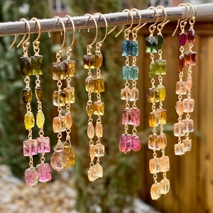 May include: A set of long, gold earrings with multiple square-shaped gemstones in shades of green, blue, pink, and yellow. The earrings are hanging from a silver bar.