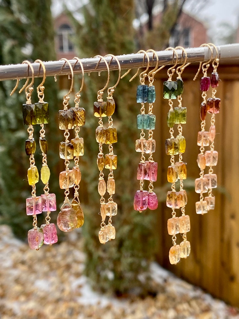 May include: A set of long, gold earrings with multiple square-shaped gemstones in shades of green, blue, pink, and yellow. The earrings are hanging from a silver bar.
