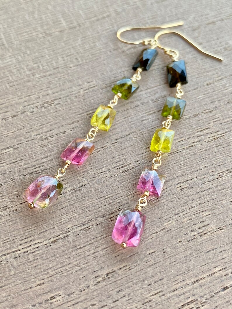 May include: A pair of gold earrings with a dangling chain of green, pink, and black gemstones. The gemstones are faceted and have a rectangular shape.