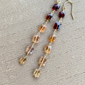 May include: Gold earrings with a graduated cascade of faceted gemstones. The earrings feature a mix of reddish-brown and golden-brown stones.