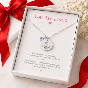 May include: A silver necklace with a round pendant engraved with a name and a heart symbol, accompanied by a small purple gemstone. The necklace is presented in a white box with the words "You Are Loved". A red ribbon is visible.
