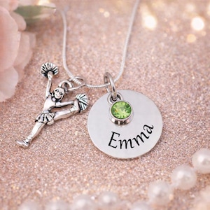 K&ouml;nnte beinhalten: Eine silberne Halskette mit zwei Anh&auml;ngern. Einer ist eine Cheerleader-Figur, der andere ein runder Anh&auml;nger mit dem Namen "Emma" und einem gr&uuml;nen Edelstein. Die Kette liegt auf einer glitzernden rosa Oberfl&auml;che.
