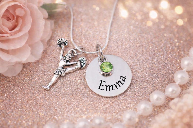 K&ouml;nnte beinhalten: Eine silberne Halskette mit zwei Anh&auml;ngern. Einer ist eine Cheerleader-Figur, der andere ein runder Anh&auml;nger mit dem Namen "Emma" und einem gr&uuml;nen Edelstein. Die Kette liegt auf einer glitzernden rosa Oberfl&auml;che.