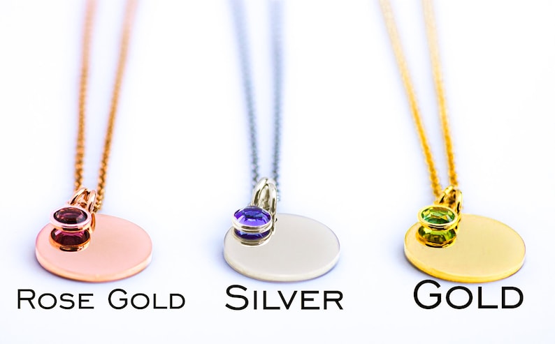 May include: Three pendant necklaces in rose gold, silver, and gold. Each necklace features a round pendant with a gemstone accent. The rose gold pendant has a red gemstone, the silver has a purple gemstone, and the gold has a green gemstone. The text "ROSE GOLD", "SILVER", and "GOLD" are below each necklace.