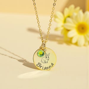 May include: A gold-colored pendant necklace featuring a round charm with a bunny illustration and the name "Brianna" in script. A green gemstone is set above the charm. The necklace is on a gold chain.