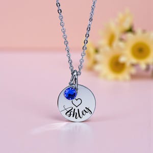 May include: A silver-toned necklace with a round pendant engraved with the name "Ashley" and a heart symbol. A blue gemstone is set above the pendant. The necklace is displayed against a soft pink background.