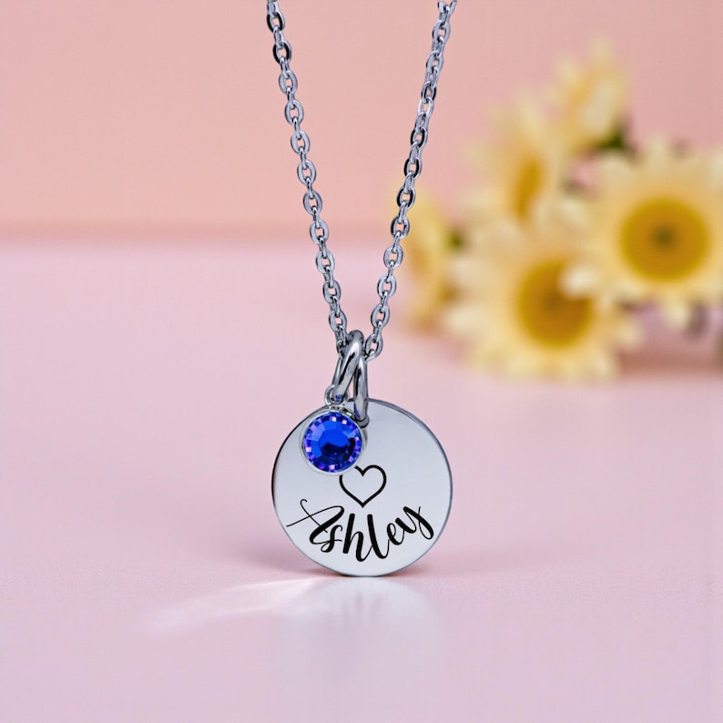 May include: A silver-toned necklace with a round pendant engraved with the name "Ashley" and a heart symbol. A blue gemstone is set above the pendant. The necklace is displayed against a soft pink background.