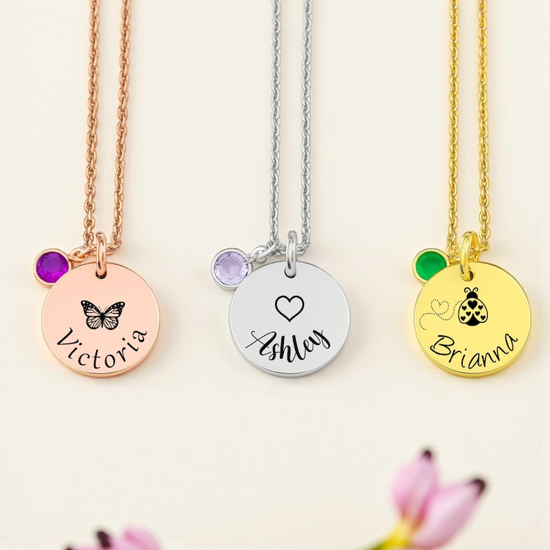 May include: Three personalized pendant necklaces. One is rose gold with a butterfly and the name "Victoria". Another is silver with a heart and the name "Ashley". The third is gold with a ladybug and the name "Brianna". Each has a gemstone.