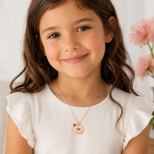 May include: A young girl wearing a rose gold necklace with a round pendant. The pendant features a butterfly design, the name "Victoria", and a small purple gemstone. The girl is wearing a white top.