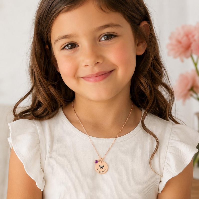 May include: A young girl wearing a rose gold necklace with a round pendant. The pendant features a butterfly design, the name "Victoria", and a small purple gemstone. The girl is wearing a white top.