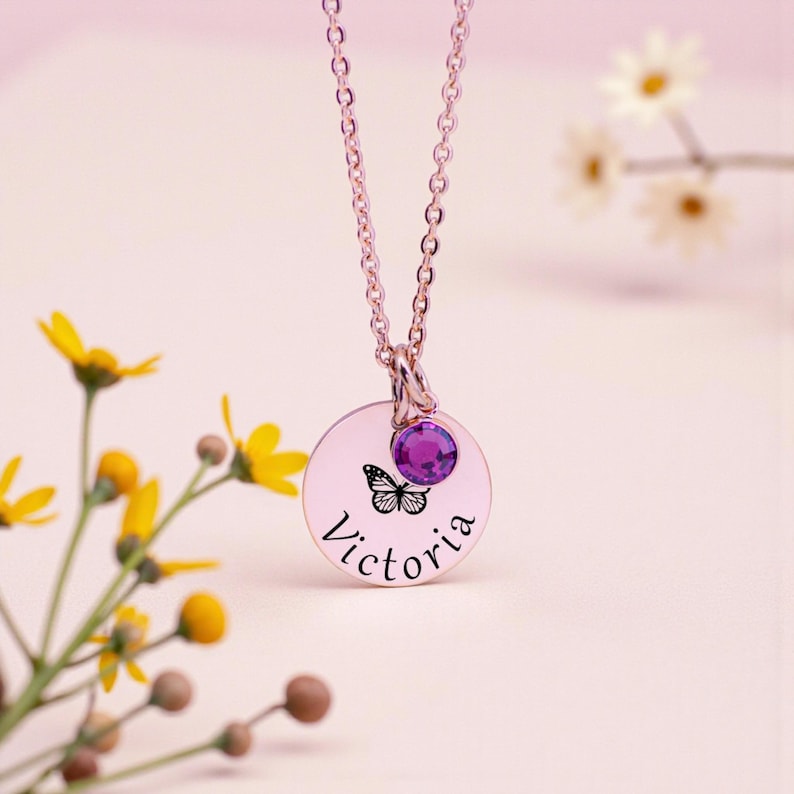May include: A rose gold necklace with a round pendant featuring a butterfly and the name "Victoria" in script. A purple gemstone is set above the pendant. The necklace is displayed against a soft pink background with yellow flowers.