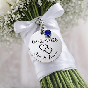 May include: A white bridal bouquet with a personalized charm. The charm is round, silver-toned, and features a blue gemstone, the date 02-21-2026, two hearts, and the names "Jon & Aimie". The bouquet is tied with a white ribbon.