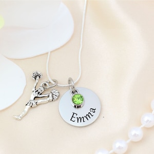K&ouml;nnte beinhalten: Silberkette mit einem Cheerleader-Anh&auml;nger und einem runden Anh&auml;nger. Der Anh&auml;nger hat einen gr&uuml;nen Edelstein und den eingravierten Namen "Emma". Die Kette wird auf einer satinartigen Oberfl&auml;che mit einer Blume und einer Perlenkette pr&auml;sentiert.