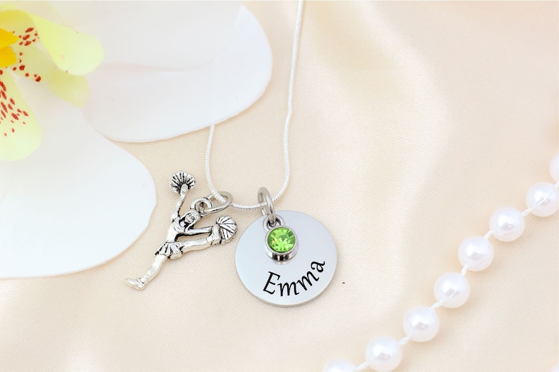 K&ouml;nnte beinhalten: Silberkette mit einem Cheerleader-Anh&auml;nger und einem runden Anh&auml;nger. Der Anh&auml;nger hat einen gr&uuml;nen Edelstein und den eingravierten Namen "Emma". Die Kette wird auf einer satinartigen Oberfl&auml;che mit einer Blume und einer Perlenkette pr&auml;sentiert.