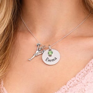 K&ouml;nnte beinhalten: Silberkette mit drei Anh&auml;ngern: einer Cheerleader-Figur, einem runden Anh&auml;nger mit dem eingravierten Namen "Emma" und einem gr&uuml;nen Edelstein. Die Kette wird am Hals einer Person getragen.