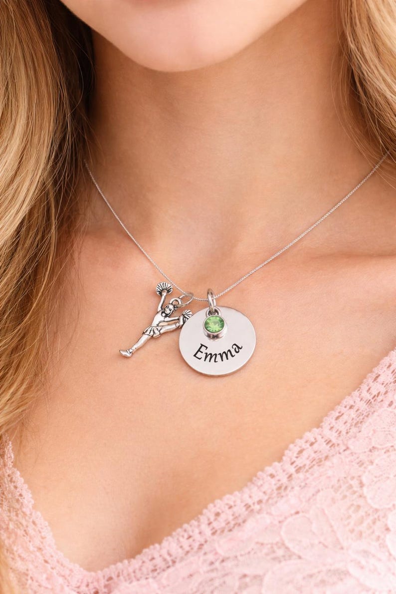 K&ouml;nnte beinhalten: Silberkette mit drei Anh&auml;ngern: einer Cheerleader-Figur, einem runden Anh&auml;nger mit dem eingravierten Namen "Emma" und einem gr&uuml;nen Edelstein. Die Kette wird am Hals einer Person getragen.