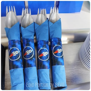 May include: Four silver plastic forks wrapped in blue napkins with a space-themed design featuring a rocket ship and stars. The napkins have the text "Epic Parties by REVO" printed on them.