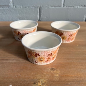 May include: Three round ceramic ramekins featuring a cream-coloured interior and an exterior decorated with a repeating pattern of orange, brown, and yellow flowers and foliage. Each small bowl has a smooth rim and a slightly tapered base design.
