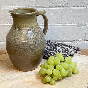 May include: A handmade ceramic jug in shades of brown and olive green, with a handle. It sits on a wooden board, alongside a bunch of green grapes and a patterned cloth. The jug is ideal for serving drinks.