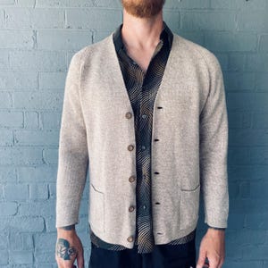 May include: A light beige cardigan sweater with a V-neck and button closure. The sweater has two front pockets and is worn over a patterned button-down shirt. The background is a blue brick wall.
