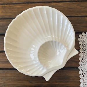 May include: A glossy off-white ceramic dish shaped like a large scallop shell. The surface features deep, radiating fluted ridges that extend from the base to the scalloped outer edge, creating a textured, fan-like appearance with a smooth finish.