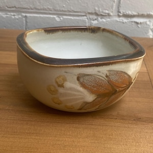May include: A square ceramic bowl with a cream exterior and a dark brown rim. The bowl has a floral design in brown and tan. The interior is white, and it sits on a wooden surface. The bowl is suitable for serving.