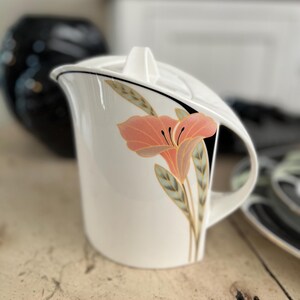 May include: A white ceramic jug with a lid, featuring a stylised peach-coloured flower graphic with green leaves and a black accent line. The jug has a curved handle and a tapered spout, showcasing a smooth, glossy finish on the exterior surface.